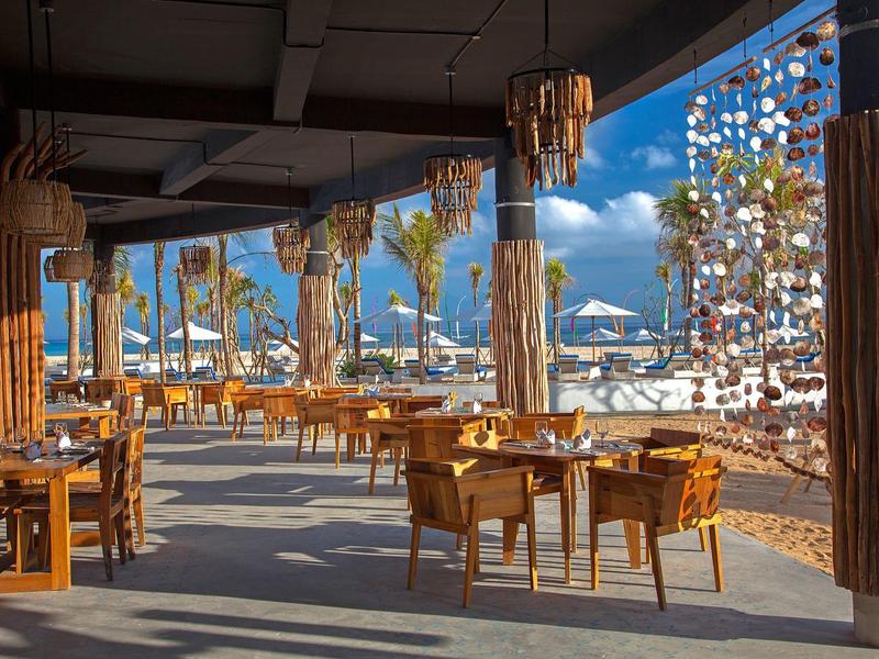 Ristorante all'aperto con mobili in legno con vista sulla spiaggia e sul mare in una giornata di sole.
