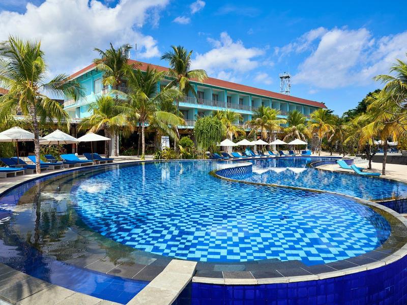 Piscina rotonda con piastrelle blu davanti a un hotel con palme e ombrelloni.