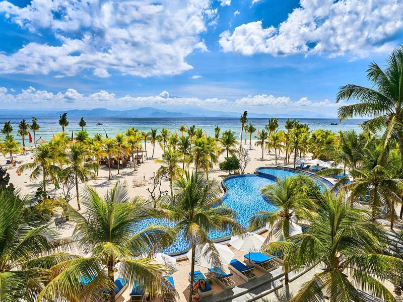 Resort sulla spiaggia con palme, piscina e lettini sotto un cielo nuvoloso.