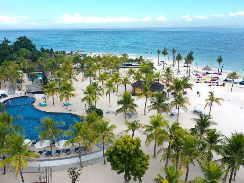 Complesso alberghiero con più piscine, palme e spiaggia di sabbia bianca sul mare.