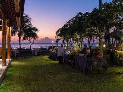 Wieczorna scena w ogrodzie z stolikami, kwiatami i palmami nad wodą z górami w tle.