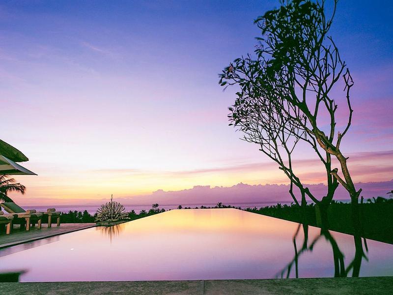 Basen infinity o zachodzie słońca z odbijającą się wodą i drzewem na pierwszym planie.