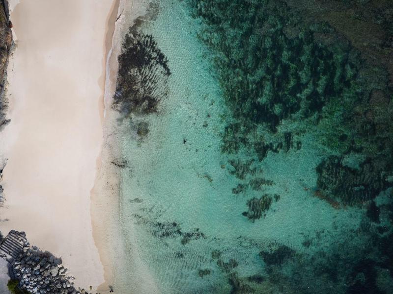 Aerial view of a white sandy beach with clear turquoise water and dark rocks.