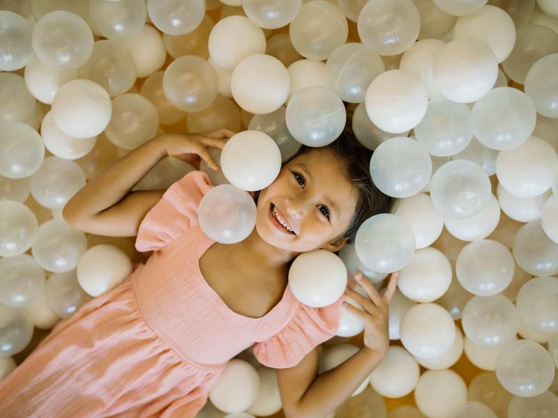 A smiling girl lies in a ball pit filled with white balls.