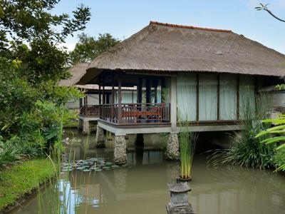 Une maison traditionnelle sur pilotis au-dessus d'un étang, entourée de plantes vertes et d'arbres.