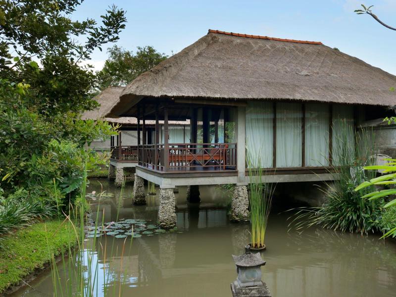 Une maison traditionnelle sur pilotis au-dessus d'un étang, entourée de plantes vertes et d'arbres.