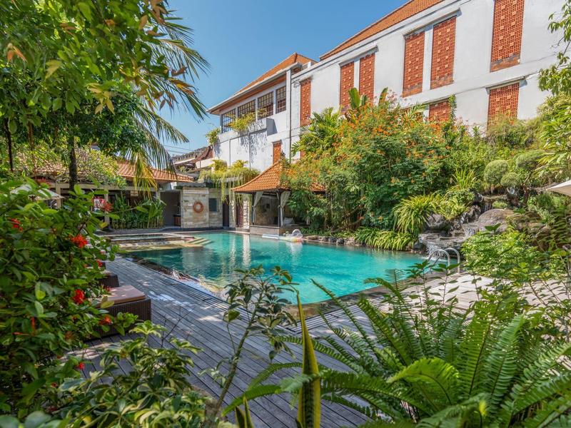 Ogród hotelowy z basenem, otoczony bujną tropikalną roślinnością i wielopiętrowym budynkiem.