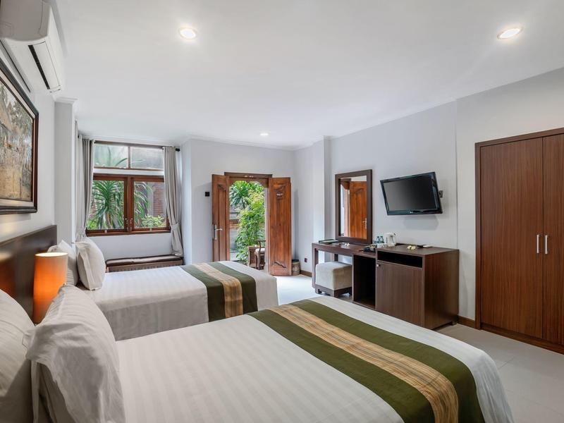 Jasny pokój hotelowy z dwoma łóżkami, drewnianymi szafami, telewizorem i balkonem z widokiem na zieleń.