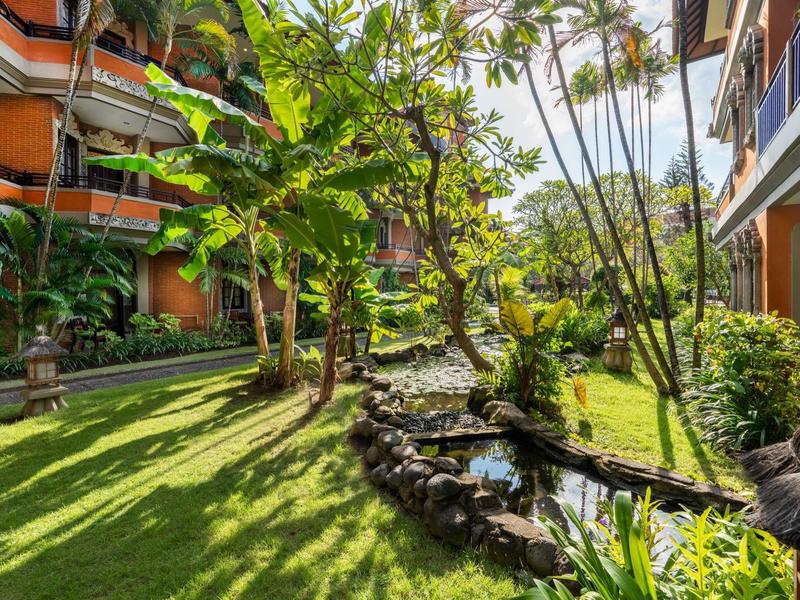 Zielony ogród hotelowy ze ścieżkami, strumieniem i tropikalnymi roślinami między budynkami.