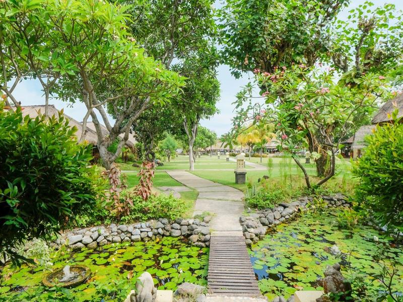 Ein Holzsteg führt durch einen grünen Garten mit Seerosen und Bäumen zu einem offenen Bereich.