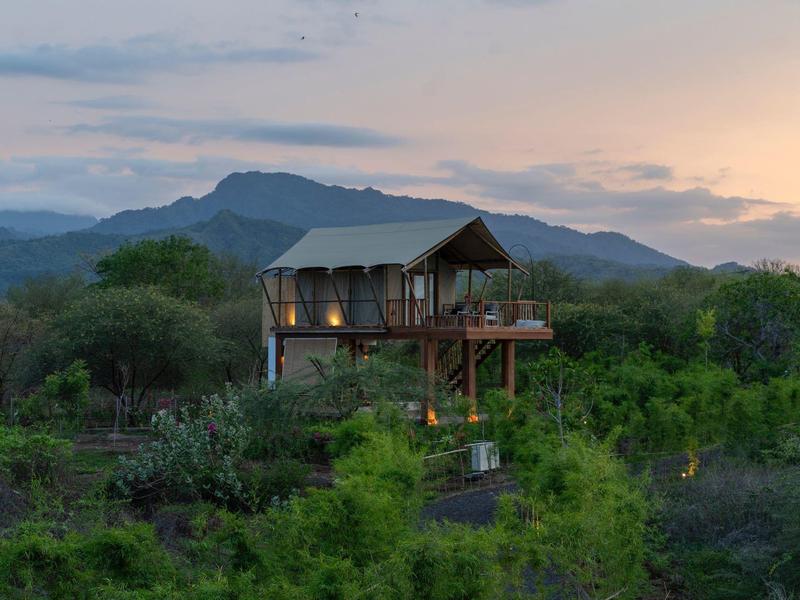 Una casa sull'albero elevata nel verde con montagne sullo sfondo al tramonto.