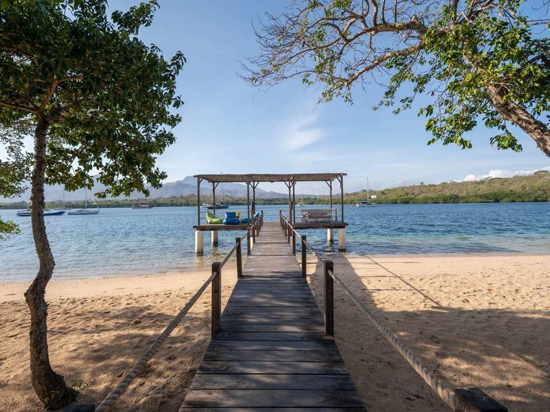 Molo di legno conduce dalla spiaggia sabbiosa a un pontile coperto sull'acqua sotto un cielo azzurro.
