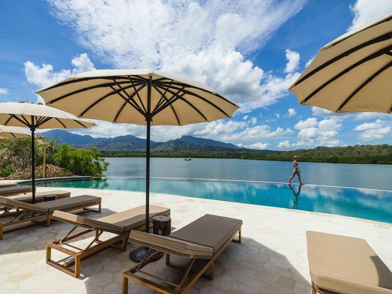 Area piscina con lettini e ombrelloni con vista su acqua e montagne.