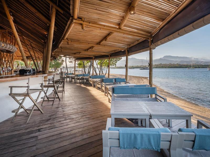Ristorante aperto sulla spiaggia con pavimento in legno e vista sul mare sotto cielo sereno.
