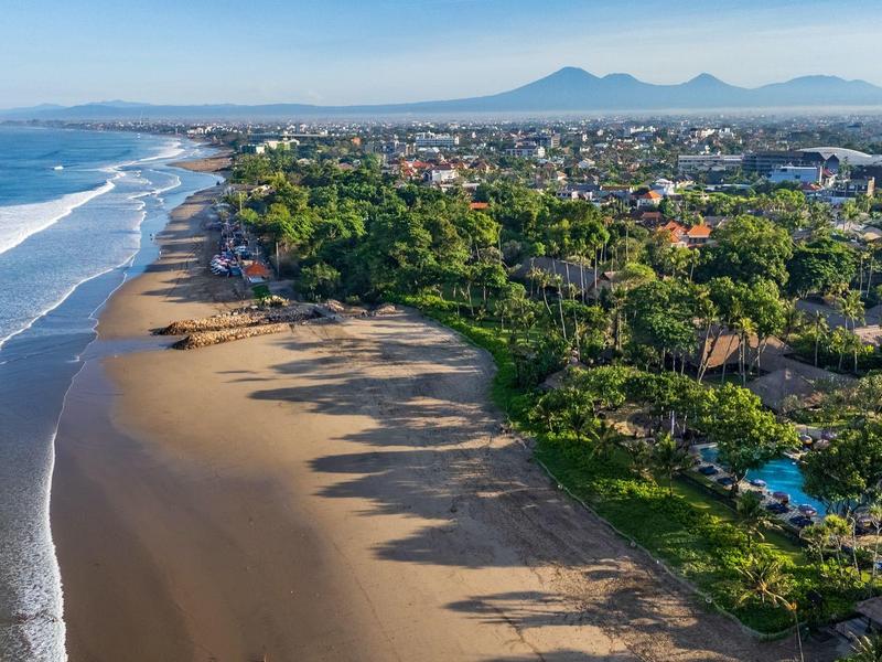 Luchtfoto van een lang zandstrand met groene kustvegetatie en een stad op de achtergrond.