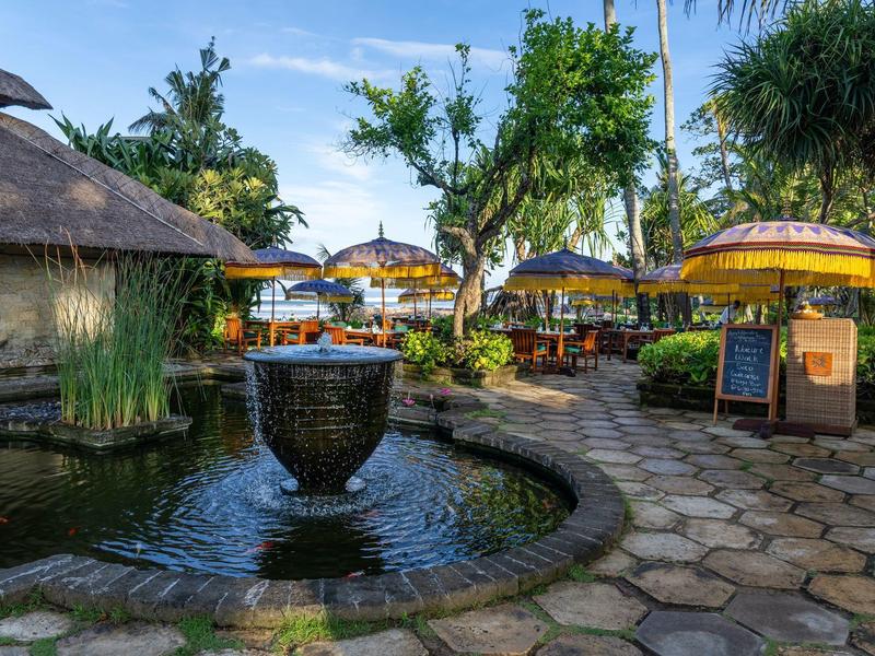 Een tropische tuin met een ronde vijver en parasols naast een restaurant.