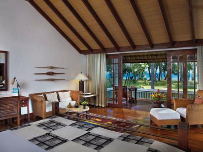 Helles Hotelzimmer mit Holzmöbeln, Sitzbereich und Blick auf einen Garten mit Palmen.