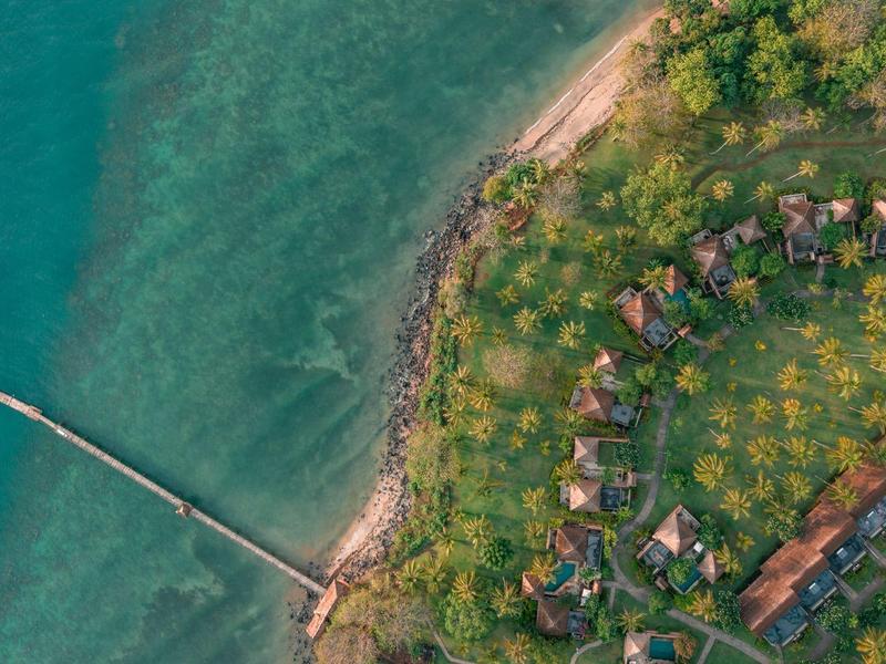 Luftaufnahme eines Meeresufers mit Steg, Strand und bewaldetem Bereich mit Gebäuden.