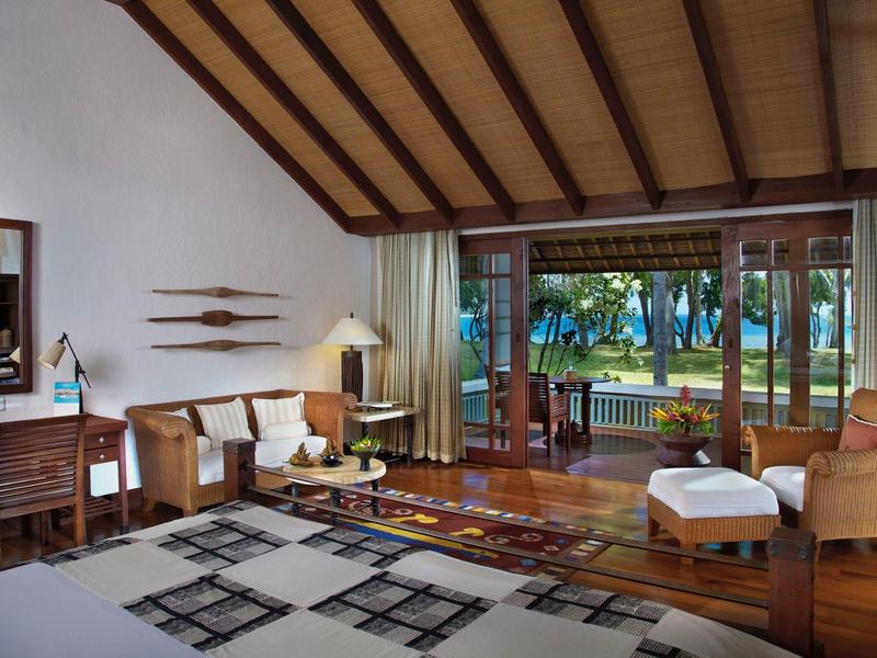 Helles Hotelzimmer mit Holzmöbeln, Sitzbereich und Blick auf einen Garten mit Palmen.