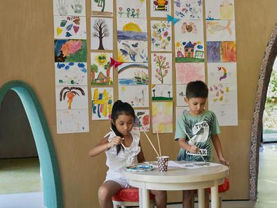 Zwei Kinder sitzen an einem runden Tisch mit Malutensilien, umgeben von an der Wand hängenden Kunstwerken.