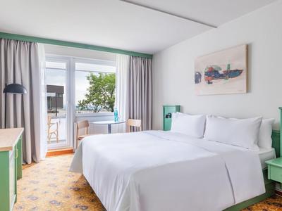 Helles Hotelzimmer mit Doppelbett, grünem Kopfteil und Balkon mit Meerblick.