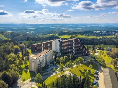 Großes Hotel in moderner Architektur umgeben von Wald und grünen Landschaften unter blauem Himmel.