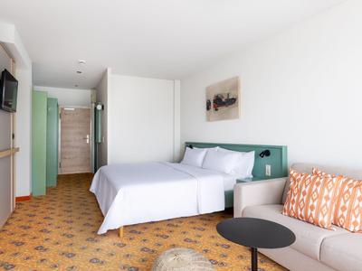 Modernes Hotelzimmer mit Doppelbett, Sofa, TV und offenem Badebereich in heller Ausstattung.