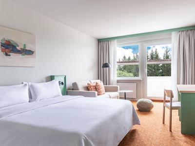 Modernes Hotelzimmer mit großem Bett, Sofa und Fenstern mit Blick ins Grüne.