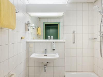 Helles Badezimmer mit weiß gefliesten Wänden, Waschbecken, Spiegel, Duschbereich und gelben Handtüchern.