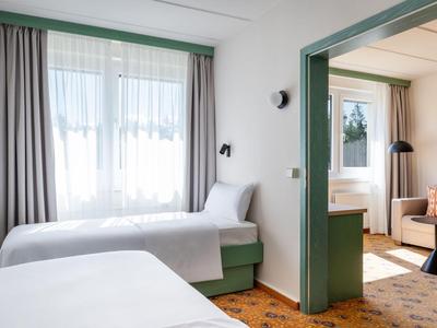 Hell eingerichtetes Hotelzimmer mit zwei Einzelbetten, Teppichboden und angrenzendem Wohnzimmer.