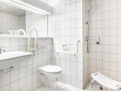 Helles, weiß gekacheltes Hotelbadezimmer mit Toilette, Waschbecken und barrierefreier Dusche mit Sitz.