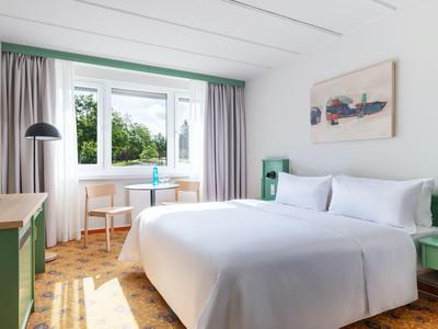 Helles Hotelzimmer mit großem Bett, grünem Mobiliar und Fenster mit Ausblick auf Bäume.