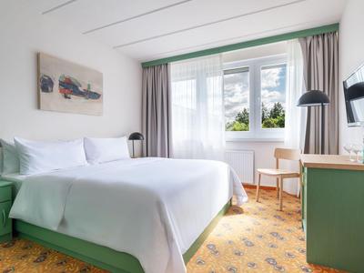 Helles Hotelzimmer mit großem Bett, grünem Mobiliar und Fenster mit Blick ins Freie.