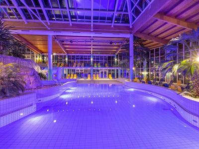 Innenpool mit blauer Beleuchtung in modernem Hotel mit Glasdach und Pflanzen.
