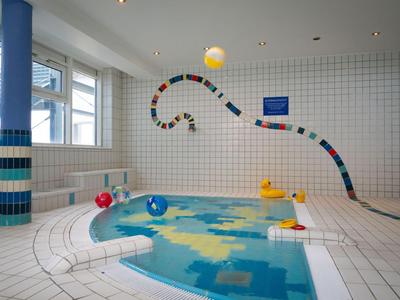 Kleines Hallenbad mit weißen Fliesen, bunten Bällen und gelben Enten als Spielzeug.
