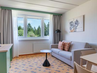 Helles Hotelzimmer mit Sofa, kleinem Tisch, TV und großem Fenster mit Blick auf Bäume.