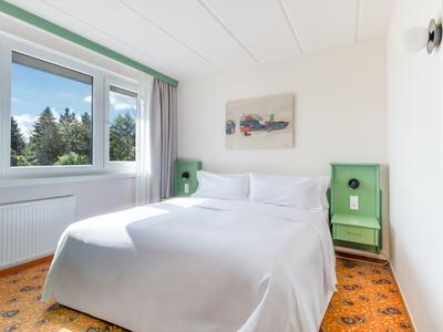 Helles, sauberes Hotelzimmer mit Doppelbett, Fenster mit Ausblick auf Bäume und grün eingerichteten Nachttischen.