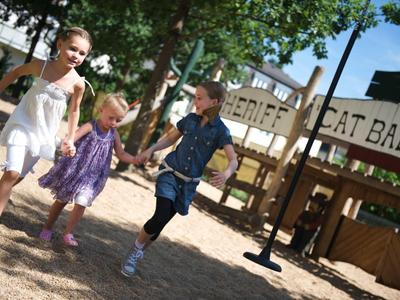 Drei Kinder in Sommerkleidung rennen lachend auf einem sonnigen Spielplatz mit Holzhäusern.