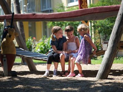Drei Kinder sitzen zusammen auf einer Holzschaukel im sonnigen Spielplatz mit Bäumen.