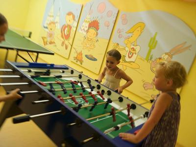 Drei Kinder spielen foosball an einem Tisch in einem Raum mit gelben Wänden und bunten Wandbildern.