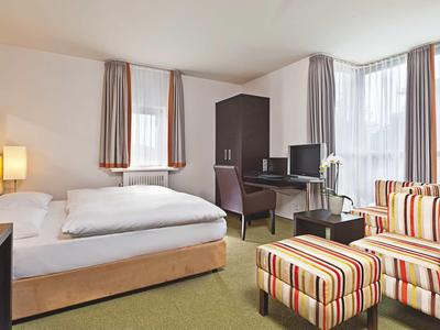 Helles Hotelzimmer mit Doppelbett, Schreibtisch, Fernseher und gestreiftem Sessel mit Hocker.