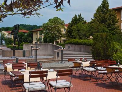 Leerer Außenbereich mit Stühlen und Tischen, Bäumen und Gebäude im Hintergrund bei Tageslicht.