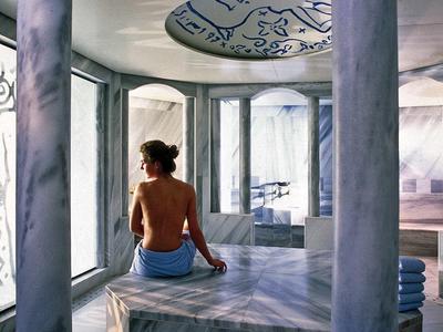 Frau sitzt in hellem, modernem Marmorraum mit Säulen und großem Fenster, trägt Handtuch um Hüfte.