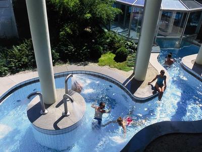 Wasserrutsche mit drei Kindern, die im klaren Wasser unter grauen Säulen rutschen, umgeben von Pflanzen.
