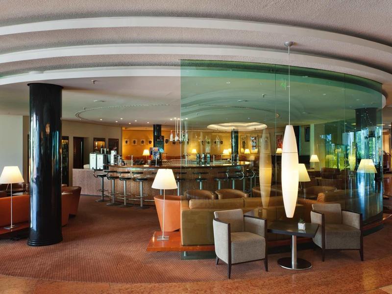 Moderner Lounge-Bereich mit abgerundeter Glaswand, braunen Sesseln, Tischen und warmem Licht.