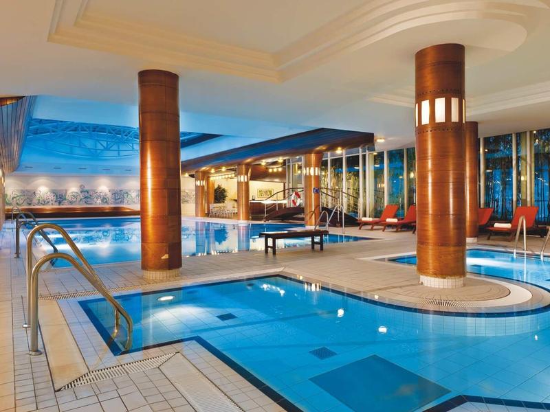 Innenbereich mit rechteckigem Pool, Holzsäulen, blauer Beleuchtung und großen Fenstern bei Tag.