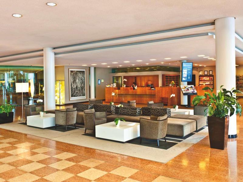 Großer, heller Hotellobbybereich mit Sitzgruppen, Pflanzen und Empfangstresen aus Holz.