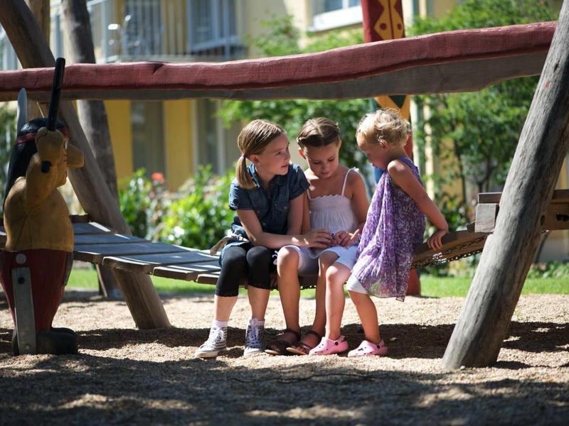 Drei Kinder sitzen zusammen auf einer Holzschaukel im sonnigen Spielplatz mit Bäumen.