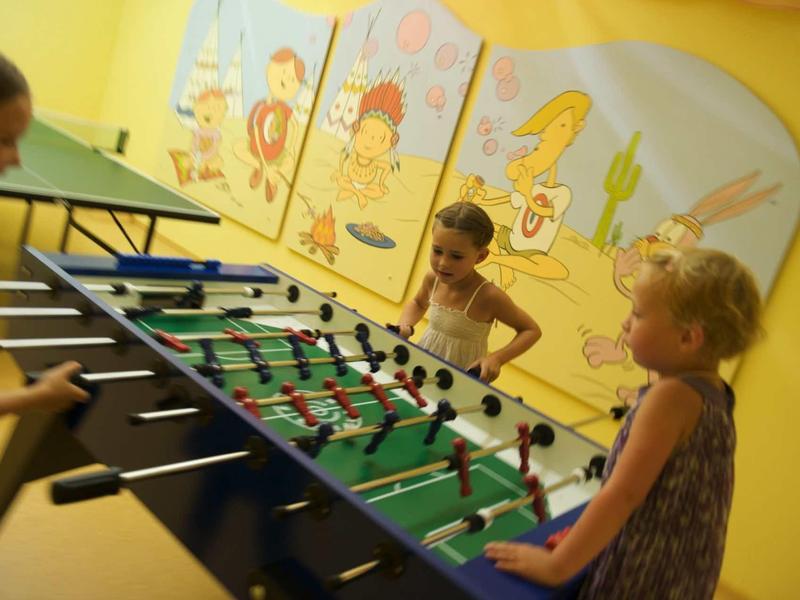 Drei Kinder spielen foosball an einem Tisch in einem Raum mit gelben Wänden und bunten Wandbildern.