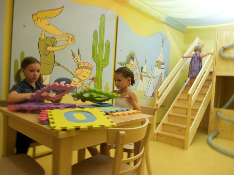 Kinder spielen an einem Holztisch mit bunten Spielmatten in einem gelben, mit Wüstenmotiven dekorierten Raum.